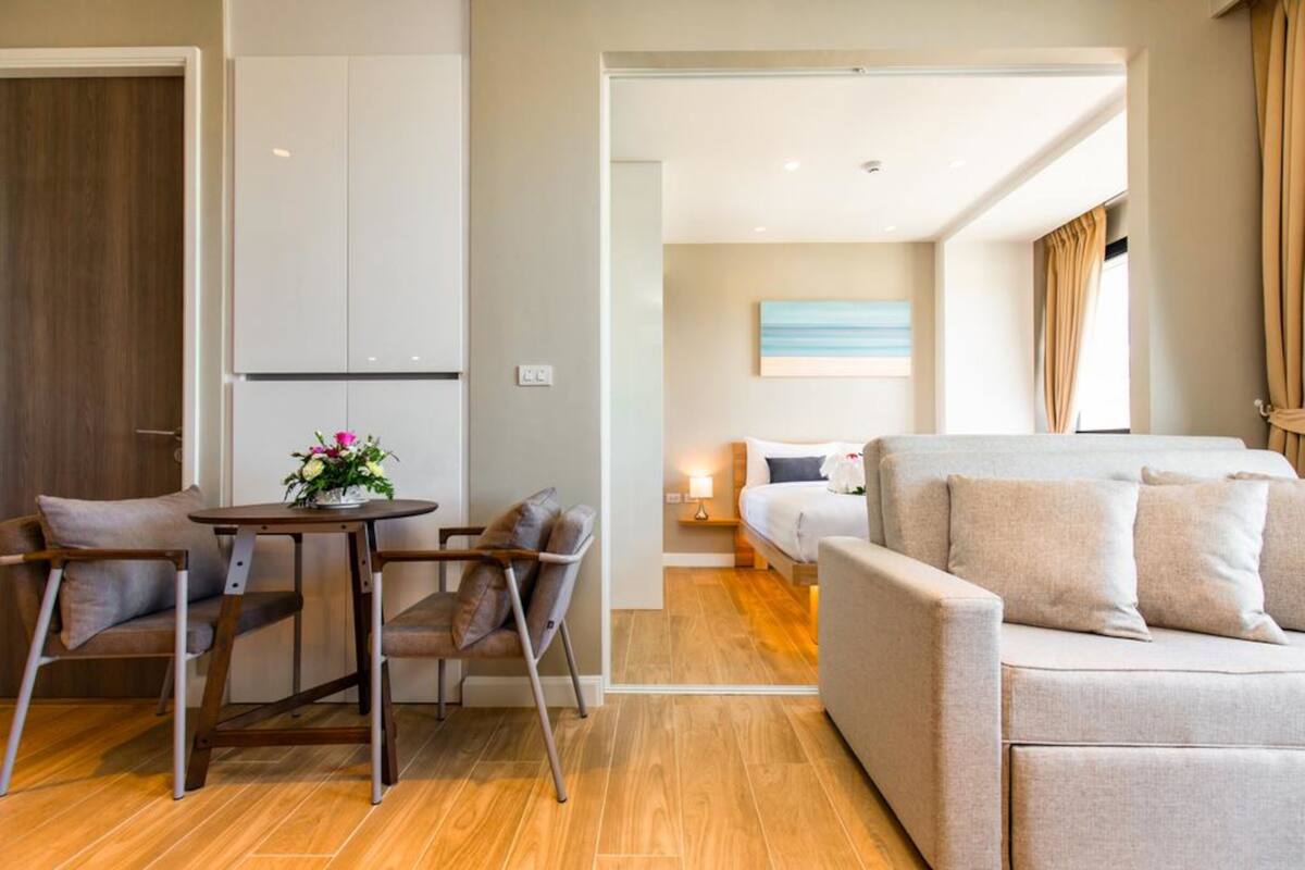 Moderni hotellihuone, jossa on puulattia, sohva, pieni pöytä maljakolla ja sänky yöpöydällä.