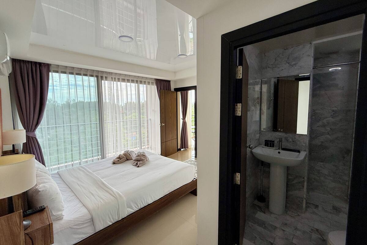 Ein modernes Hotelzimmer mit einem großen Bett, einem Nachttisch mit Lampe, einem Badezimmer mit Marmorwaschbecken und Spiegel sowie Blick durch das Fenster mit Vorhängen.