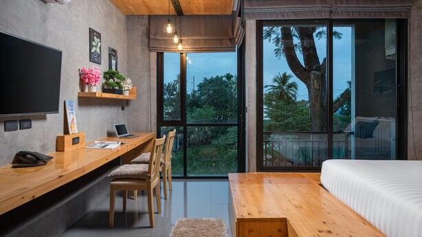A modern bedroom with a large bed, wooden desk, and a laptop. There is a TV mounted on the wall and a window with a view of trees and a palm tree.