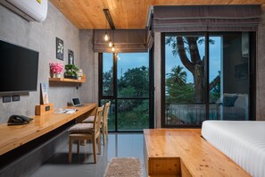 A modern bedroom with a large bed, wooden desk, and a laptop. There is a TV mounted on the wall and a window with a view of trees and a palm tree. - Relife the Windy by Villacarte (Rawai)