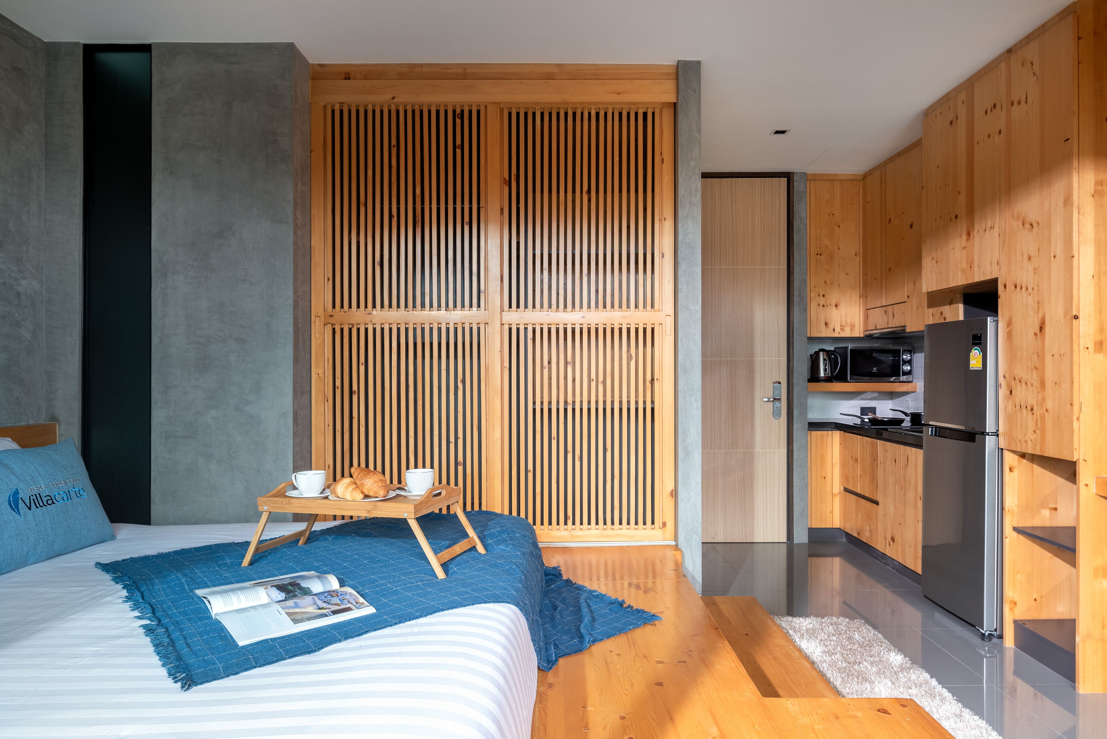 A modern hotel room with a large bed, a wooden partition, a kitchenette, and a breakfast setup on the bed.