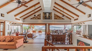 A spacious living area with a high ceiling, wooden beams, ceiling fans, and a large window offering a mountain view. -  Villa Suriya B2 (Patong)