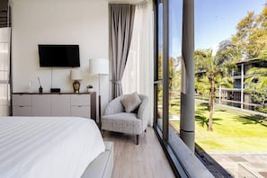 Ein Schlafzimmer mit Bett, Fernseher, Schreibtisch mit Lampe, Stuhl und Blick auf Pool und Palmen.