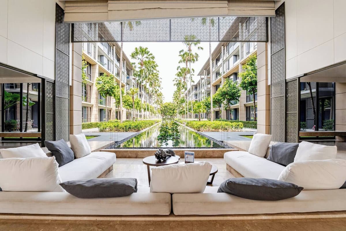 A modern hotel lobby with a central seating area, a reflective pool, and a view of the building's exterior.