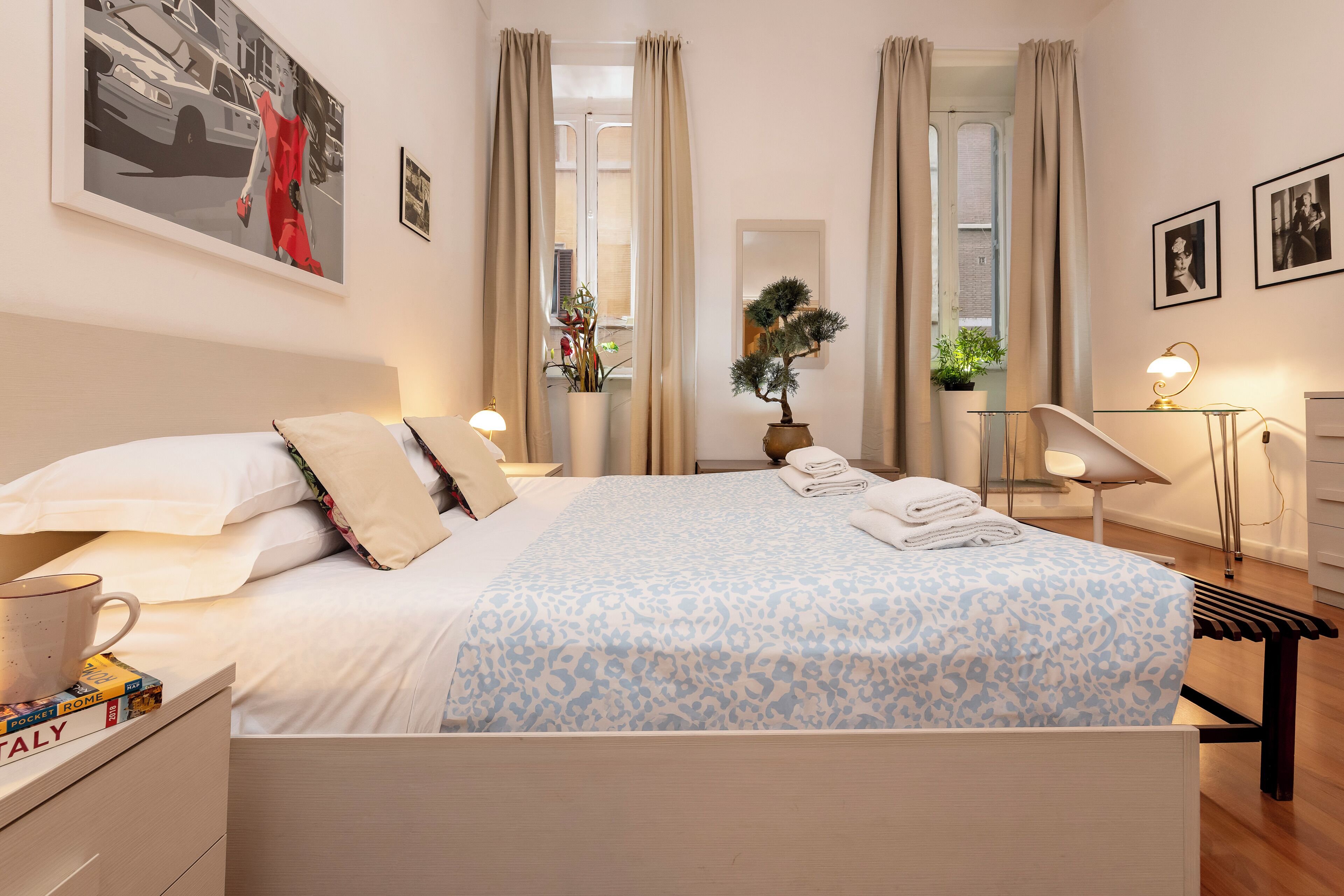 A neatly made bed with a patterned bedspread, a bedside table with a cup and books, a potted plant, and a mirror on the wall.