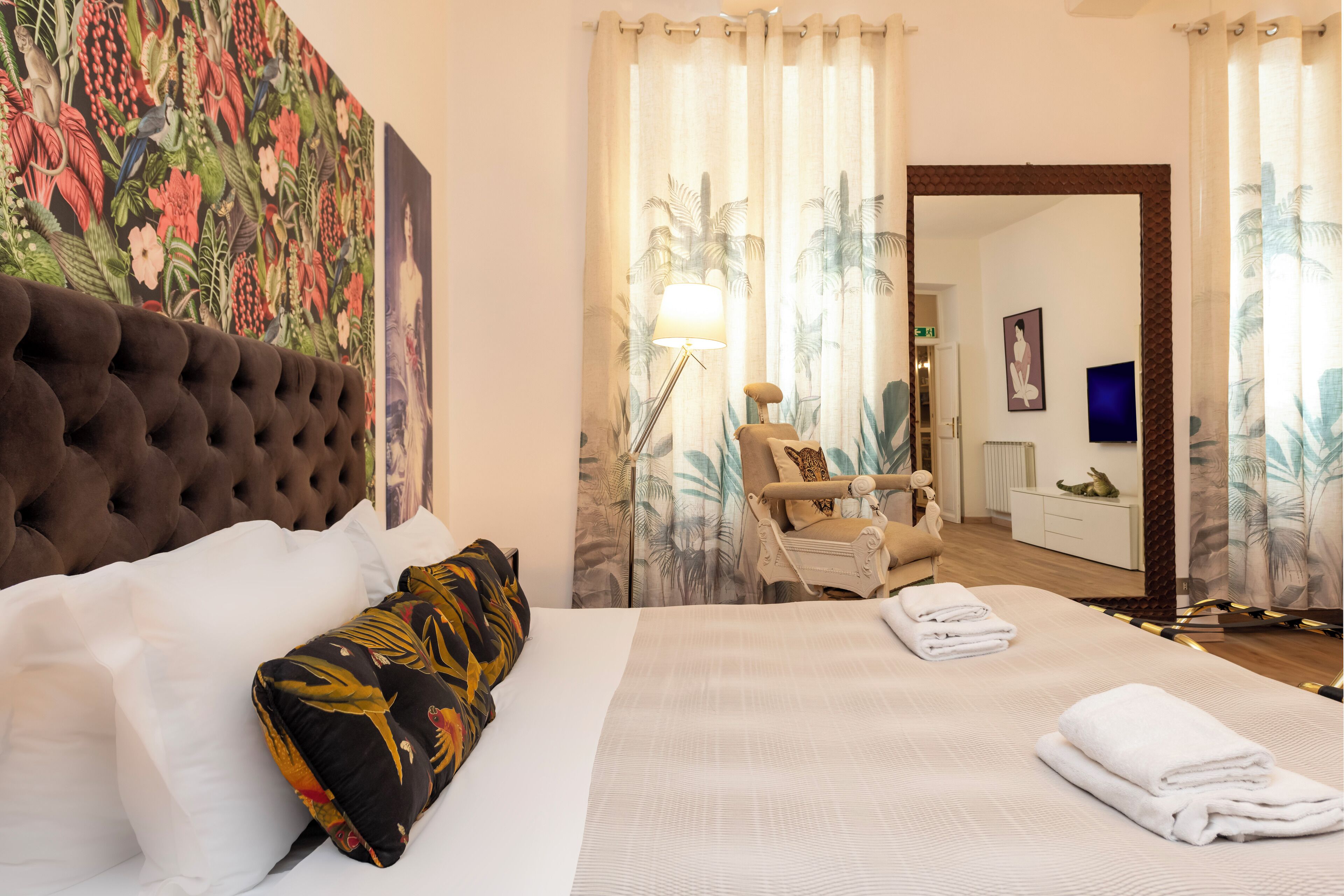 A hotel room with a tufted headboard, a large bed with white and black floral pillows, a neatly made bed with folded towels, a nightstand with a lamp, and a view of the bathroom through a curtain.