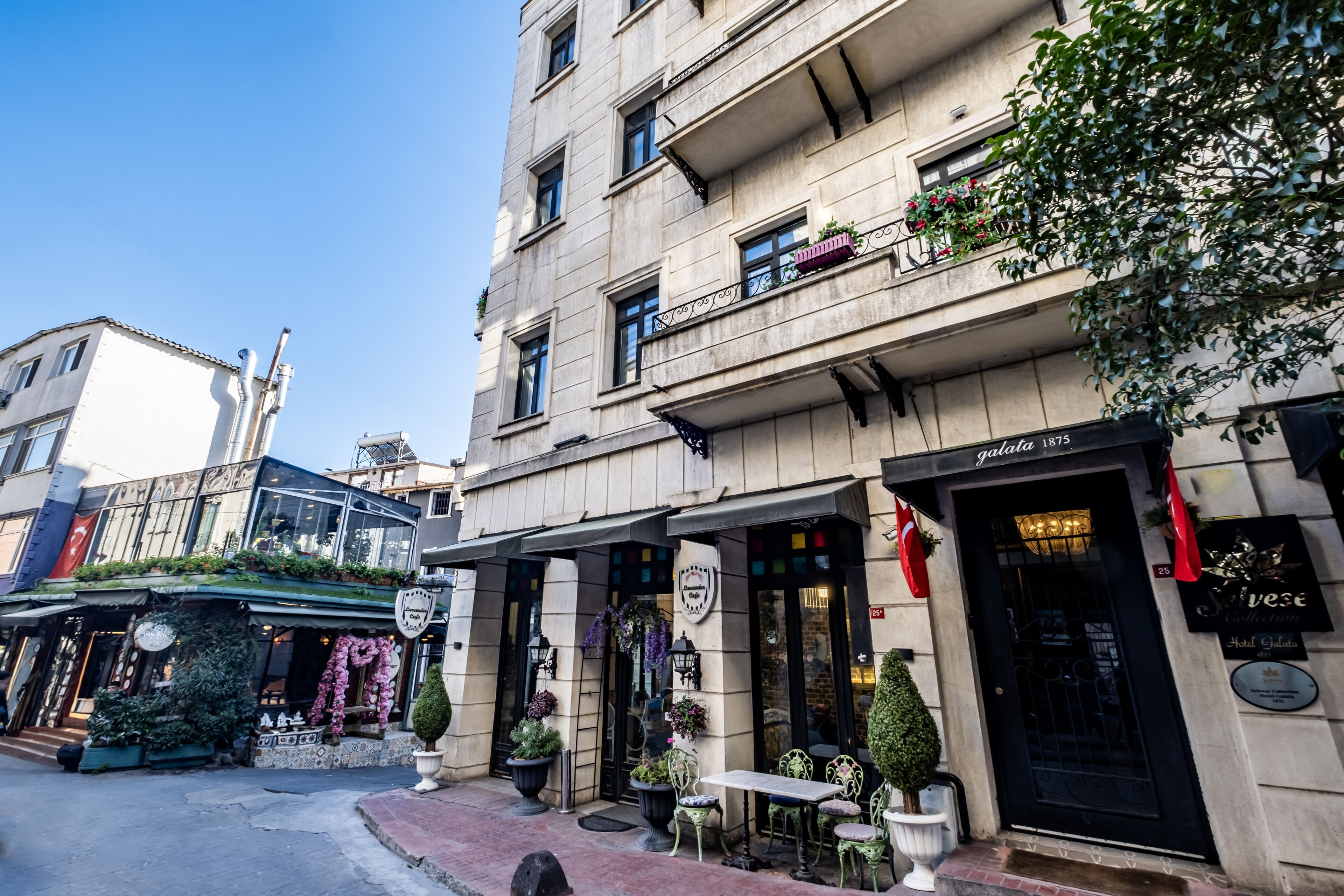 A hotel facade with a classic architectural style, featuring a black entrance door, a signboard, and outdoor seating area with potted plants.