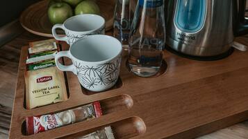 Un plateau en bois avec deux tasses blanches ornées de motifs noirs, une bouteille d’eau, une bouilloire et divers sachets de thé et de café.