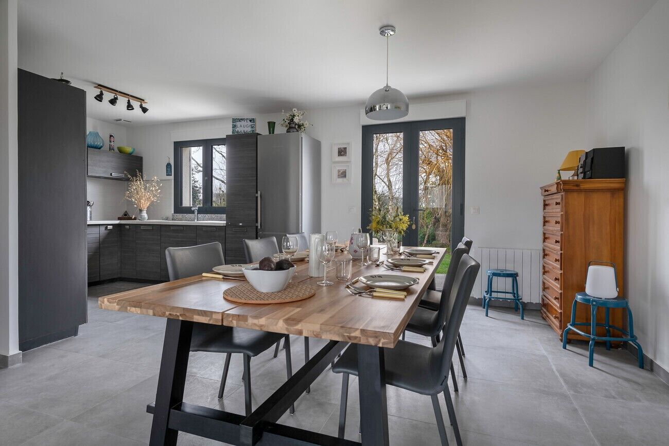 A modern kitchen with dark cabinets, a wooden dining table set for six, and a large window offering a view of the outdoors.