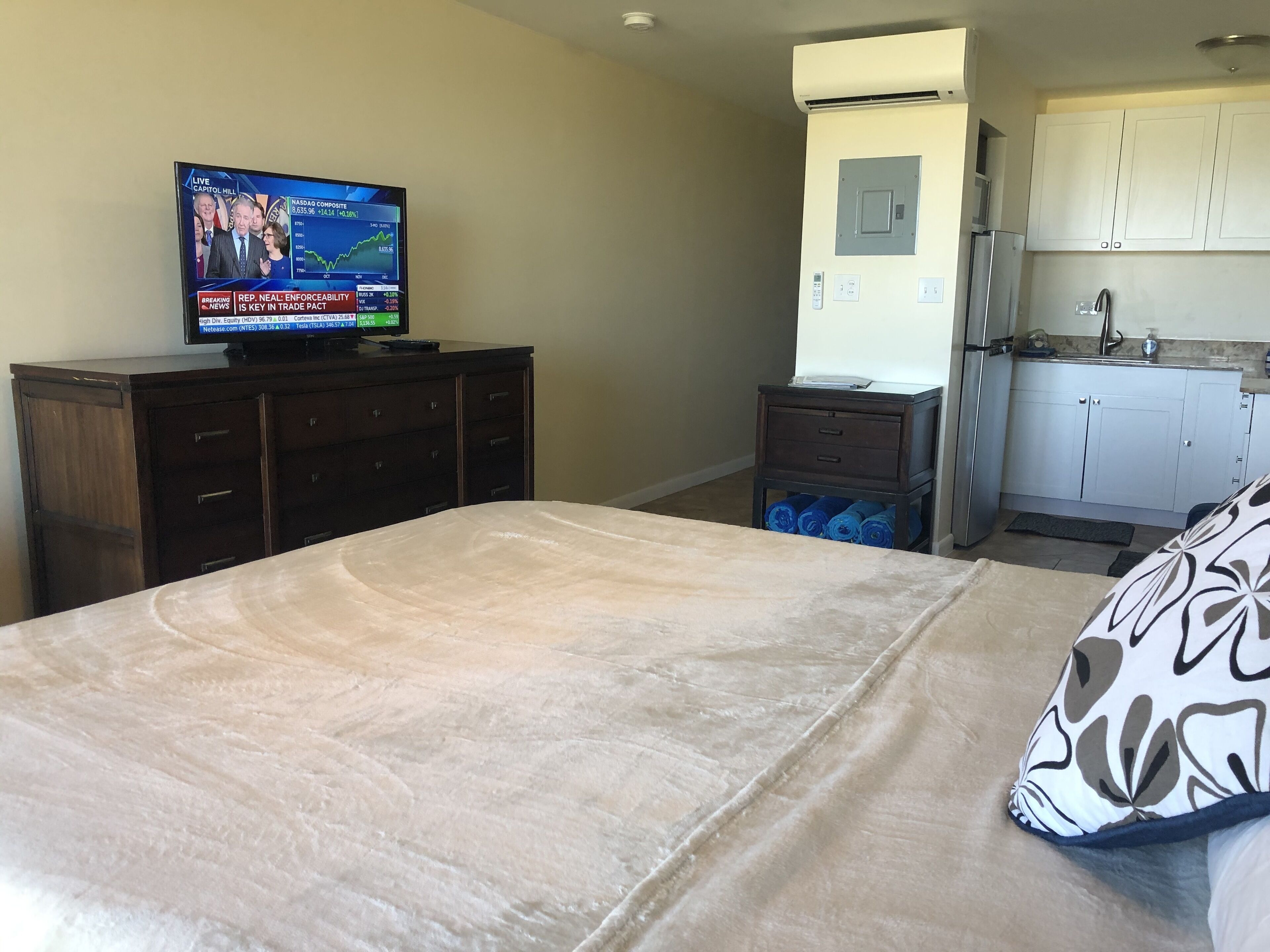 Habitación de hotel con cama, televisión mostrando un noticiero, kitchenette pequeña y baño.