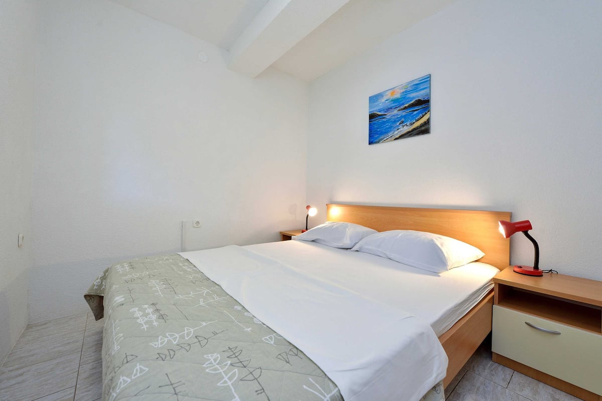 A neatly made bed with white linens, a bedside table with a red lamp, and a painting of a seascape on the wall.
