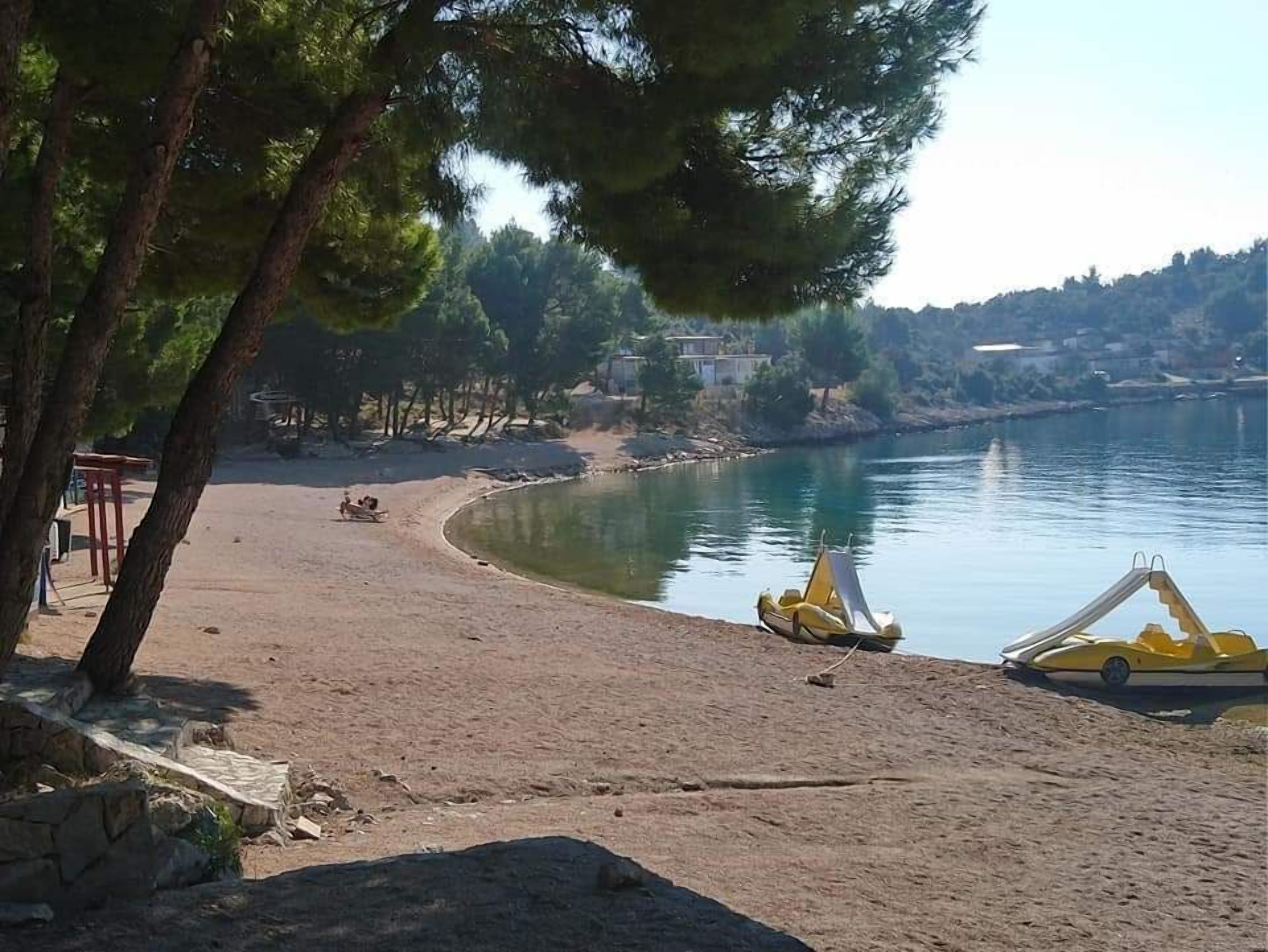 Una playa con orilla arenosa, un lago tranquilo y algunas lanchas amarillas. Se ven árboles y un edificio al fondo.
