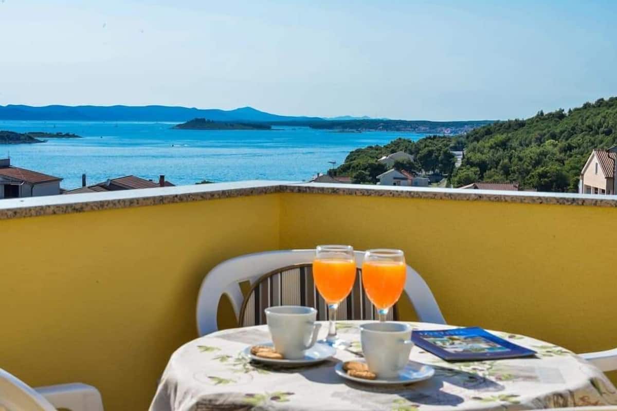 A balcony with a table set for breakfast, overlooking a coastal landscape with mountains in the distance.