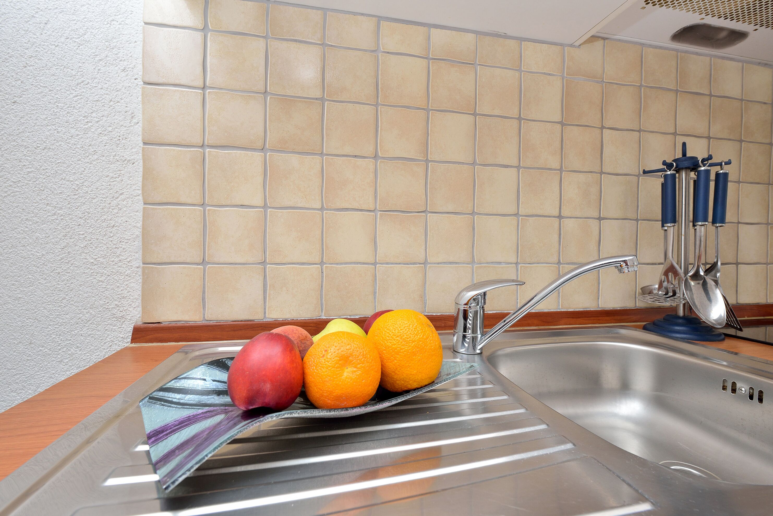 Eine Küche mit Spüle aus Edelstahl, einer Obstschale mit Äpfeln und Orangen sowie einer gefliesten Rückwand.