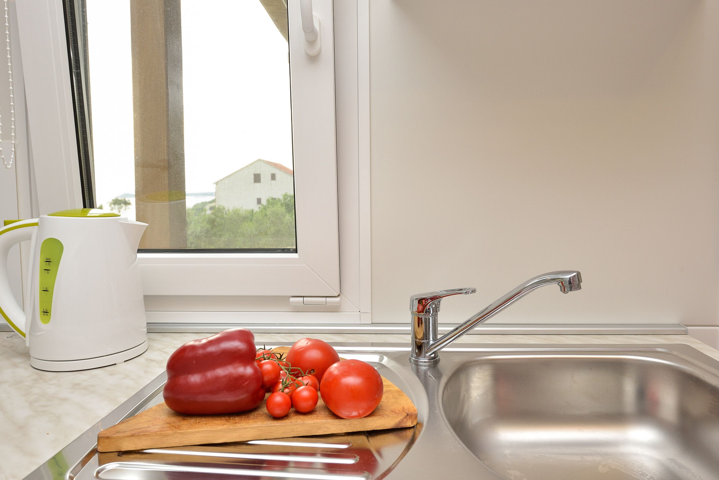 A kitchen with a stainless steel sink, a wooden cutting board with tomatoes and a red bell pepper, and a white kettle with a green handle.