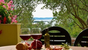 Une table avec deux verres de vin rouge, une bouteille d’huile d’olive, du pain, des tomates et une assiette de charcuterie, le tout disposé devant un paysage composé d’un lac et d’arbres.