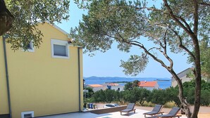 Un espace aménagé au bord de la piscine, avec des chaises longues et une table, un bâtiment jaune et une vue sur la mer.
