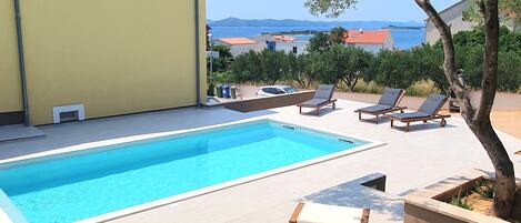 A poolside area with lounge chairs and a table, a yellow building, and a view of the sea.