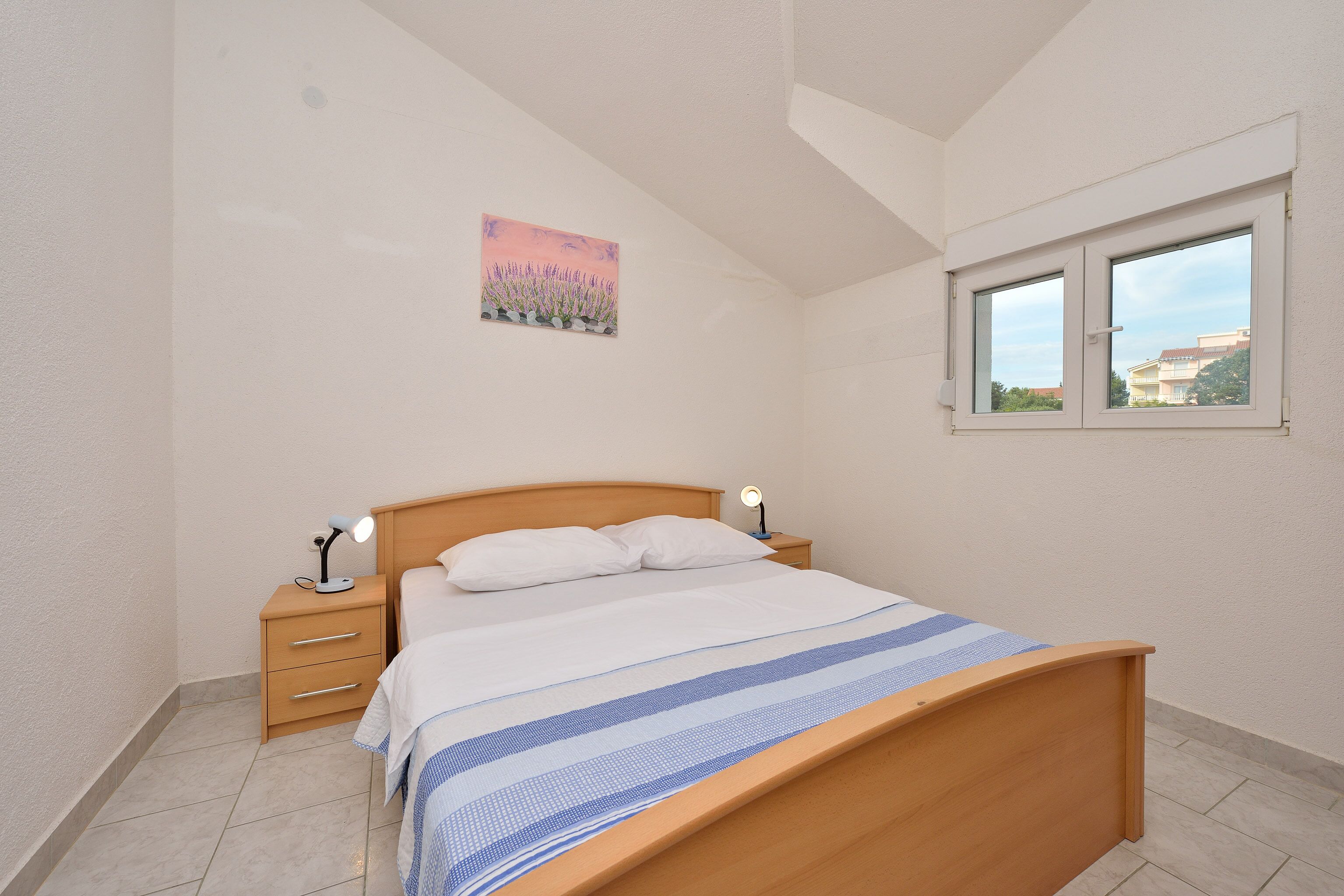 A bedroom with a wooden bed, a nightstand with a lamp, a window, and a wall-mounted picture.