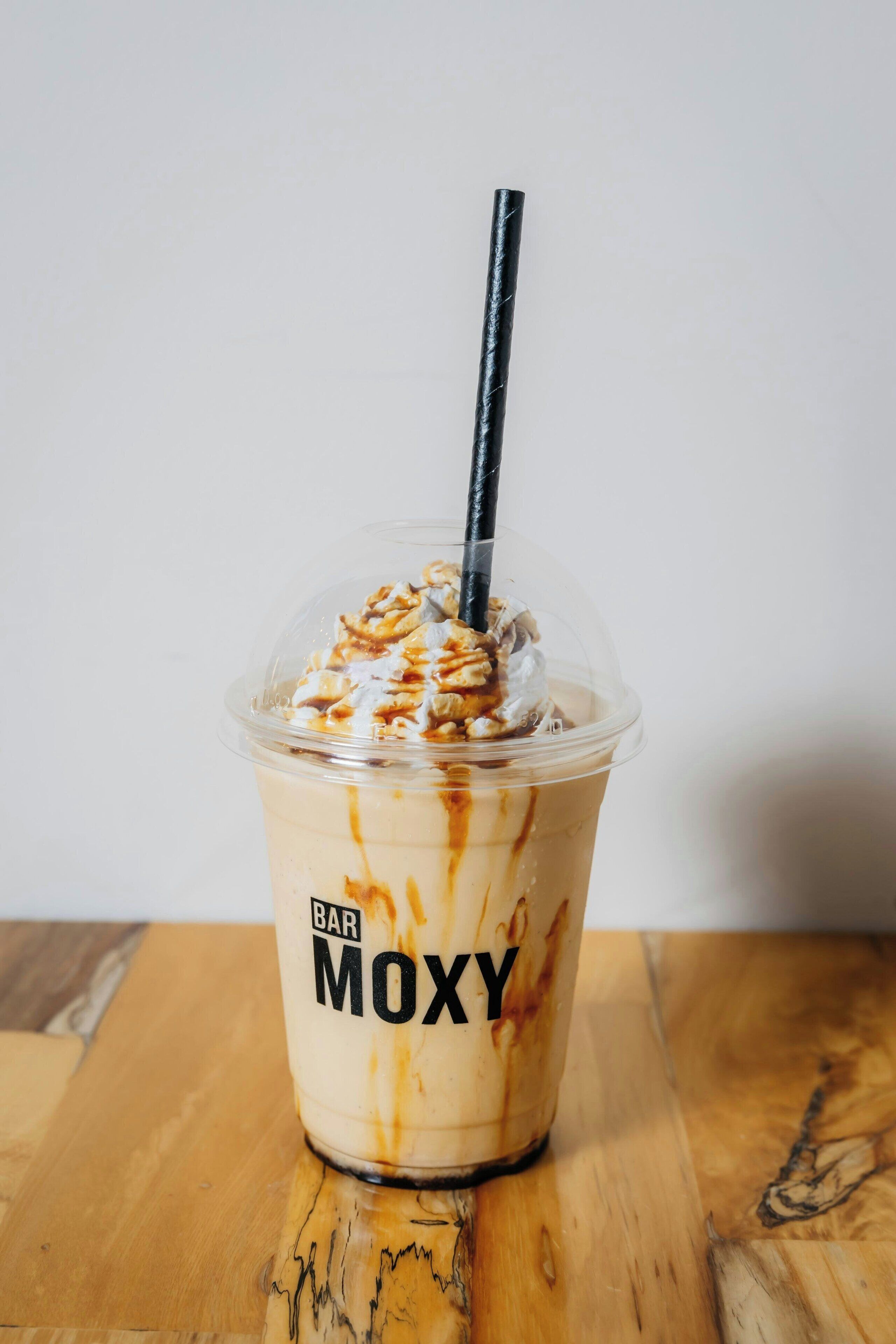 A clear plastic cup labeled "BAR MOXY" with a black straw, containing a beverage topped with whipped cream and caramel drizzle, placed on a wooden surface.