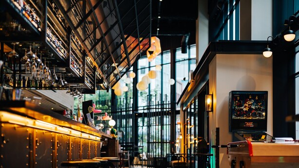 Una zona de bar con estantes llenos de botellas, luces colgantes y un amplio ventanal con luz natural.
