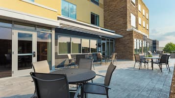 An outdoor patio with round tables and chairs, a covered seating area, and a building with large windows and a brick facade.