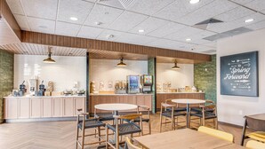 Un café moderne avec un plancher en bois, des tables rondes et des chaises. On y trouve des comptoirs avec des machines à café et un tableau affichant le menu. Une pancarte sur le mur indique : « Avancez vos montres. »