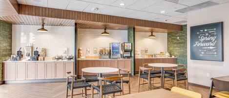 A modern cafe with wooden flooring, round tables, and chairs. There are counters with coffee machines and a menu board. A sign on the wall reads "Spring Forward."
