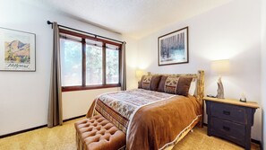 A bedroom with a large bed, a bench, a nightstand, a lamp, and two framed pictures on the walls.