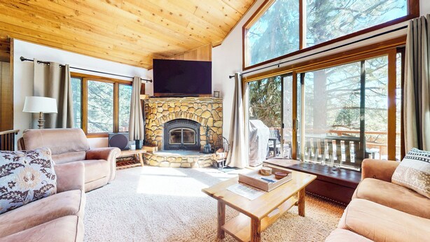 A cozy living room with a stone fireplace, large windows, and a wooden ceiling. - Updated & Large, Near Golf Course! Gas Grill & Deck. Forested Quiet Location (Mammoth Lakes)