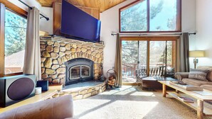 A stone fireplace, a television, a sofa, a coffee table, and a view of trees through large windows. - Updated & Large, Near Golf Course! Gas Grill & Deck. Forested Quiet Location (Mammoth Lakes)