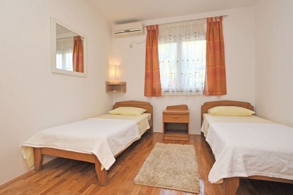 Une chambre avec deux lits simples, une petite table de chevet, une commode, un miroir, un appareil de climatisation et une fenêtre avec des rideaux.