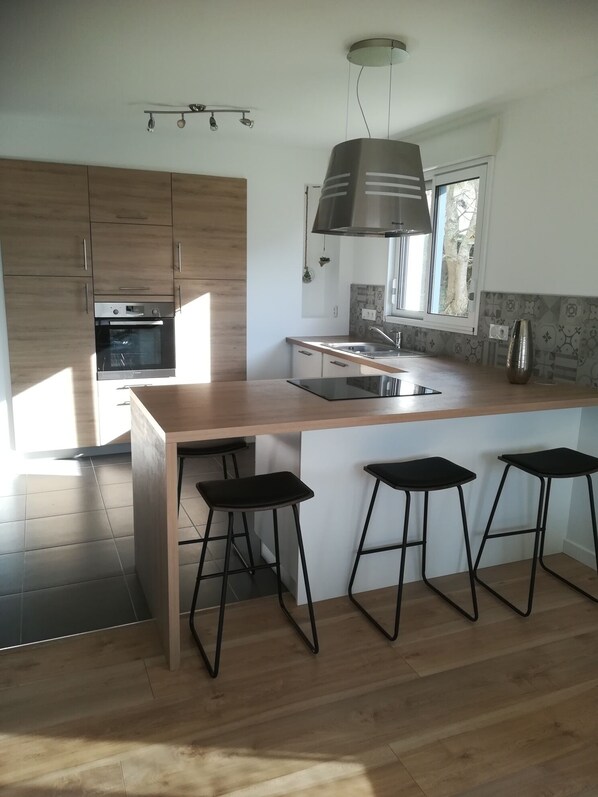 A modern kitchen with wooden cabinets, a built-in oven, a large island with stools, and a window providing natural light.