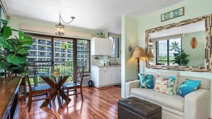 A spacious living area with a dining table, a sofa with colorful cushions, a large mirror, and a view of a residential building through the windows. - Kihei Akahi #C-520  Tastefully Updated, Corner Studio on 5th Floor, A/C (Kihei)