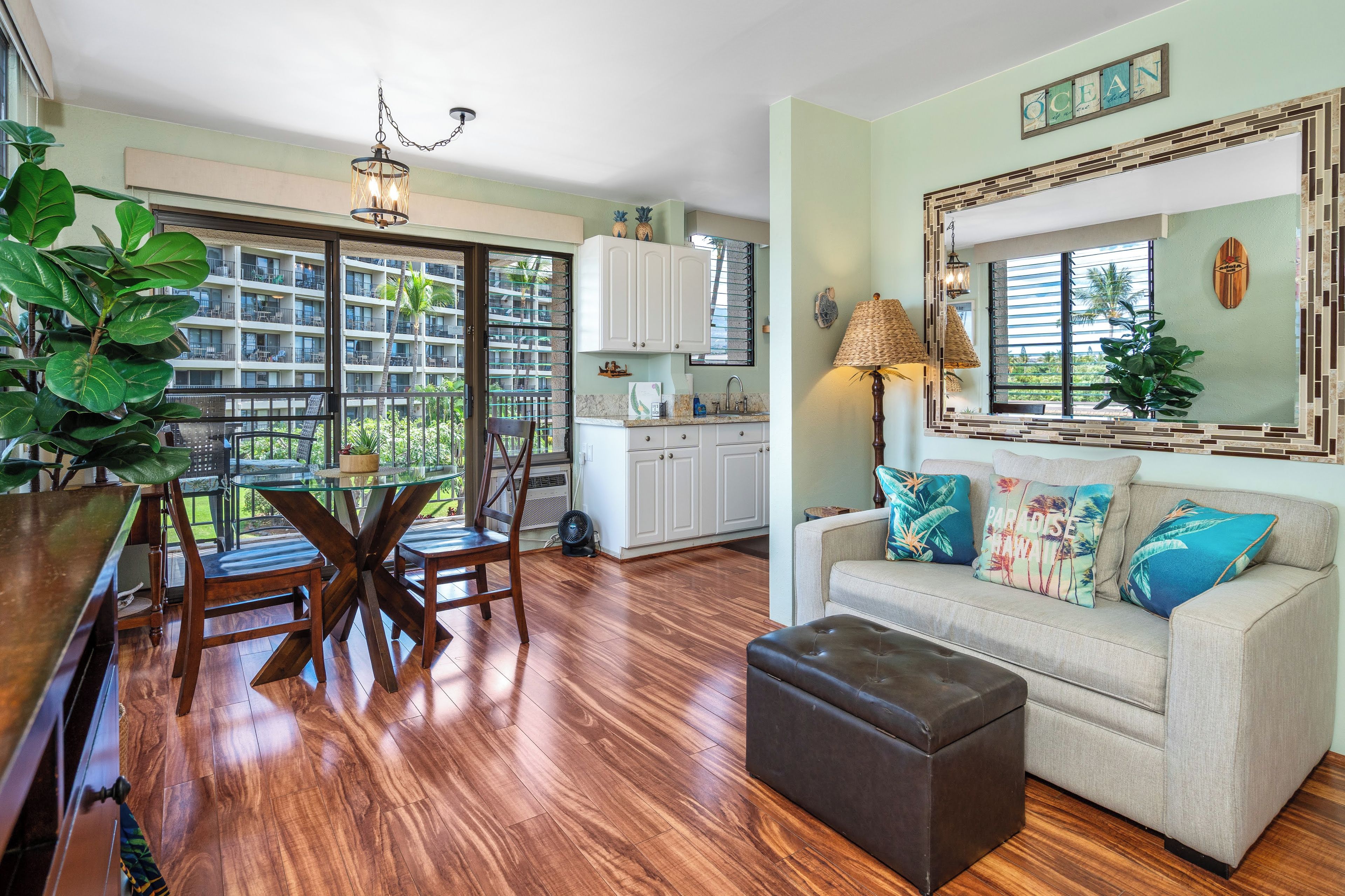 A spacious living area with a dining table, a sofa with colorful cushions, a large mirror, and a view of a residential building through the windows.