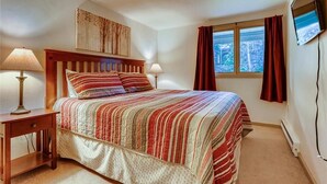 Une chambre à coucher avec un lit, deux lampes de chevet, un téléviseur fixé au mur et une fenêtre avec des rideaux.