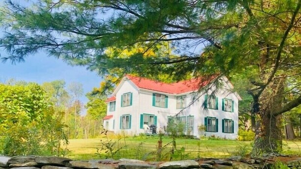 A white house with green shutters and a red roof, surrounded by trees and a stone wall. - Charming Farmhouse - quiet, private & peaceful  (Worthington)
