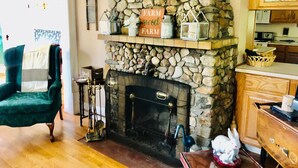 A cozy living room with a stone fireplace, a green armchair, and a kitchen area in the background. - Charming Farmhouse - quiet, private & peaceful  (Worthington)
