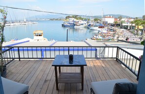 A wooden deck with a table and chairs, overlooking a marina with boats and a distant cityscape. - Capa Butik Otel (Urla)