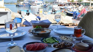 Una mesa dispuesta con alimentos para el desayuno, como huevos, aceitunas y pan, con vistas a un puerto deportivo con barcos.