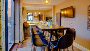 A modern kitchen with a dining area, featuring a long wooden table, bar stools, and pendant lighting. - The Station Lodge, Filey. Sleeps 12 (Filey)