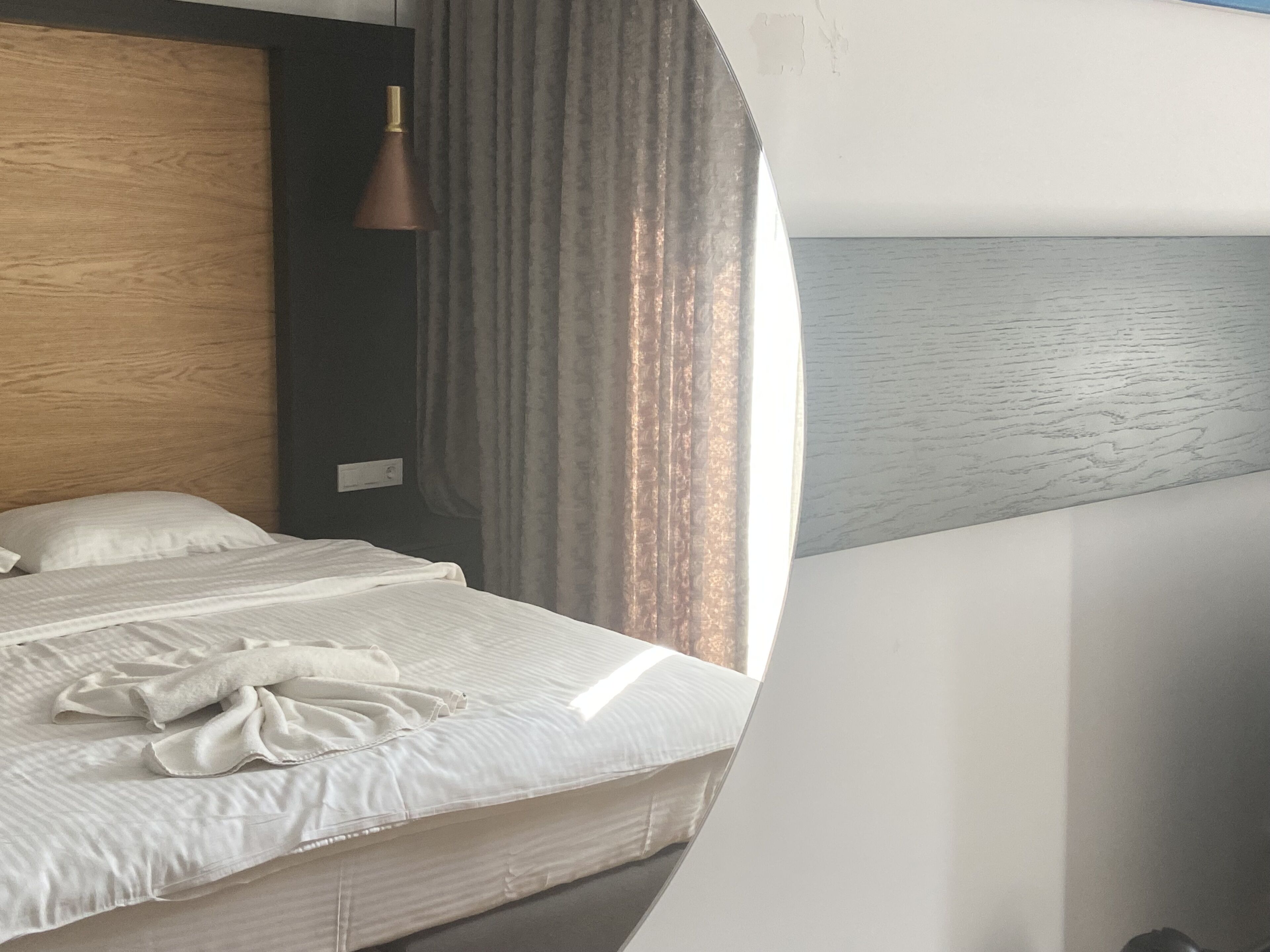 A hotel room with a bed, a wooden headboard, a pendant lamp, and a view of the sea through a window.