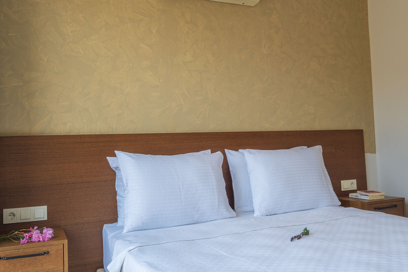 A neatly made bed with white bedding and pillows, a wooden headboard, and a bedside table with a book and a power outlet.
