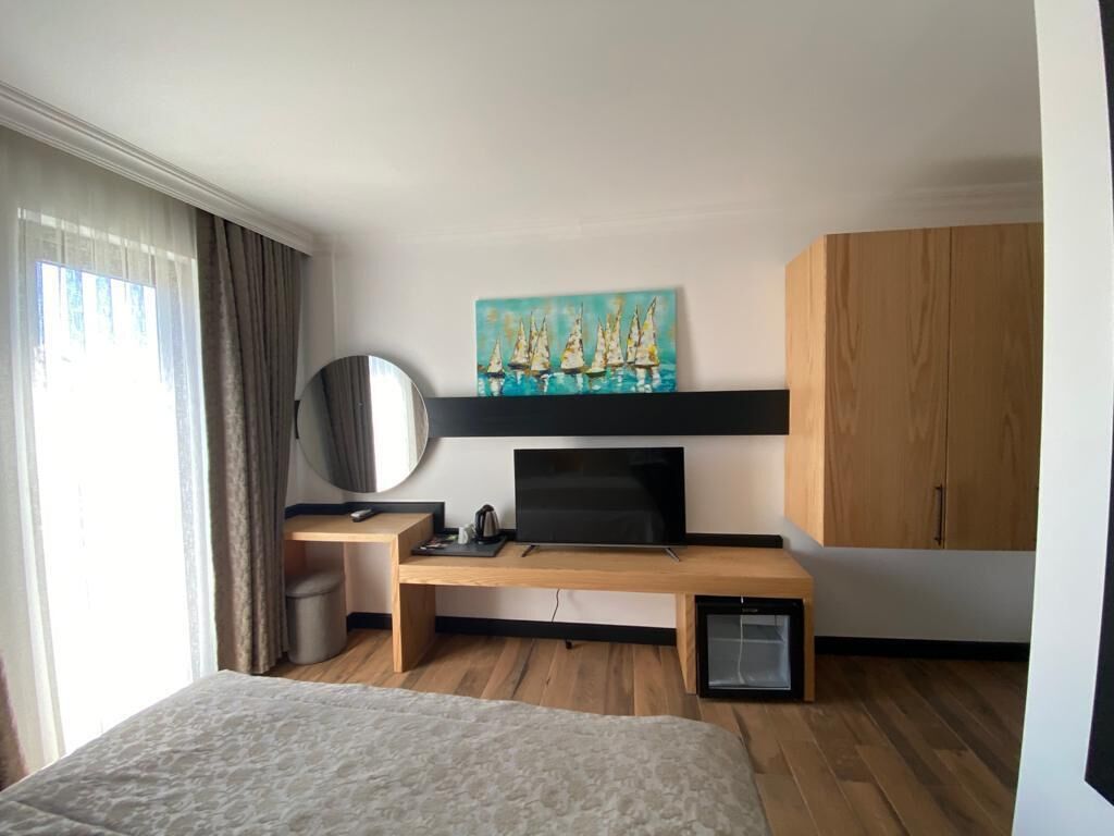 A modern hotel room with a bed, a desk with a flat-screen TV, a painting above the TV, a wooden cabinet, and a window with curtains.