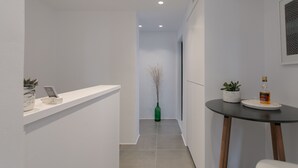 A modern hallway with a white reception desk, a small table with a bottle and glasses, and a green vase with branches. - Vorta Naxos - Adults Only (Naxos)
