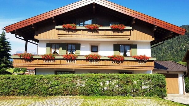 A two-story chalet with wooden balconies, each adorned with red flowers. - Breitkopf vacation apartment with south-facing terrace and mountain view (Reit im Winkl)