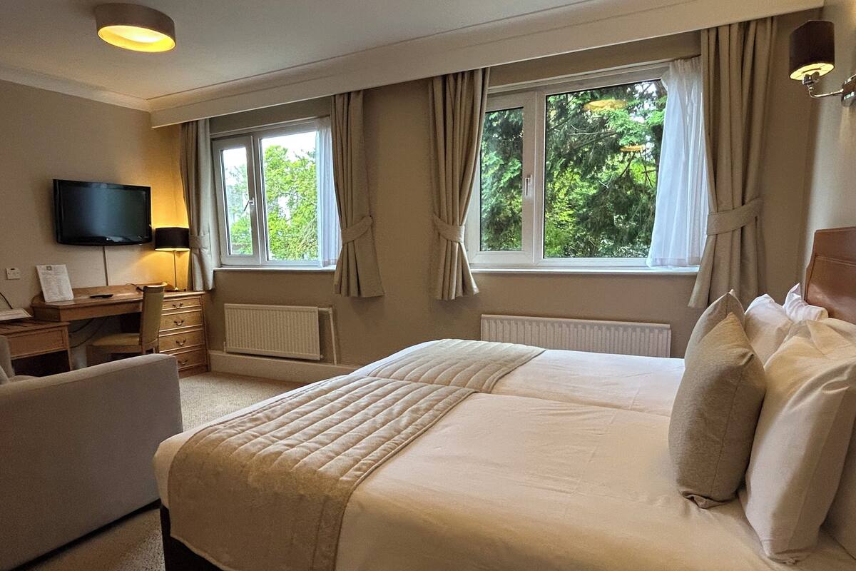 A bedroom with a bed, a desk, a chair, a television, and windows with curtains.