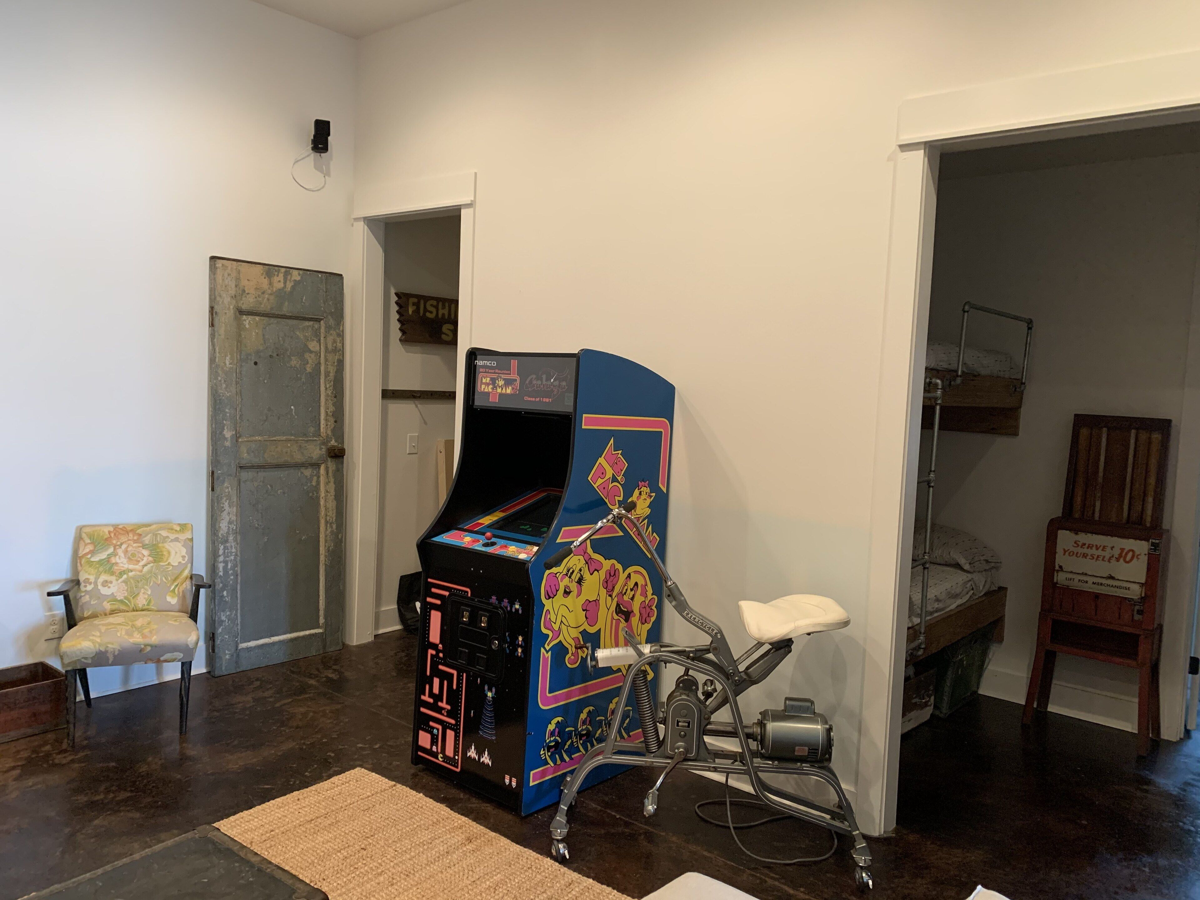 Una habitación con una máquina recreativa antigua, una silla, una cama con literas y un letrero en la pared.