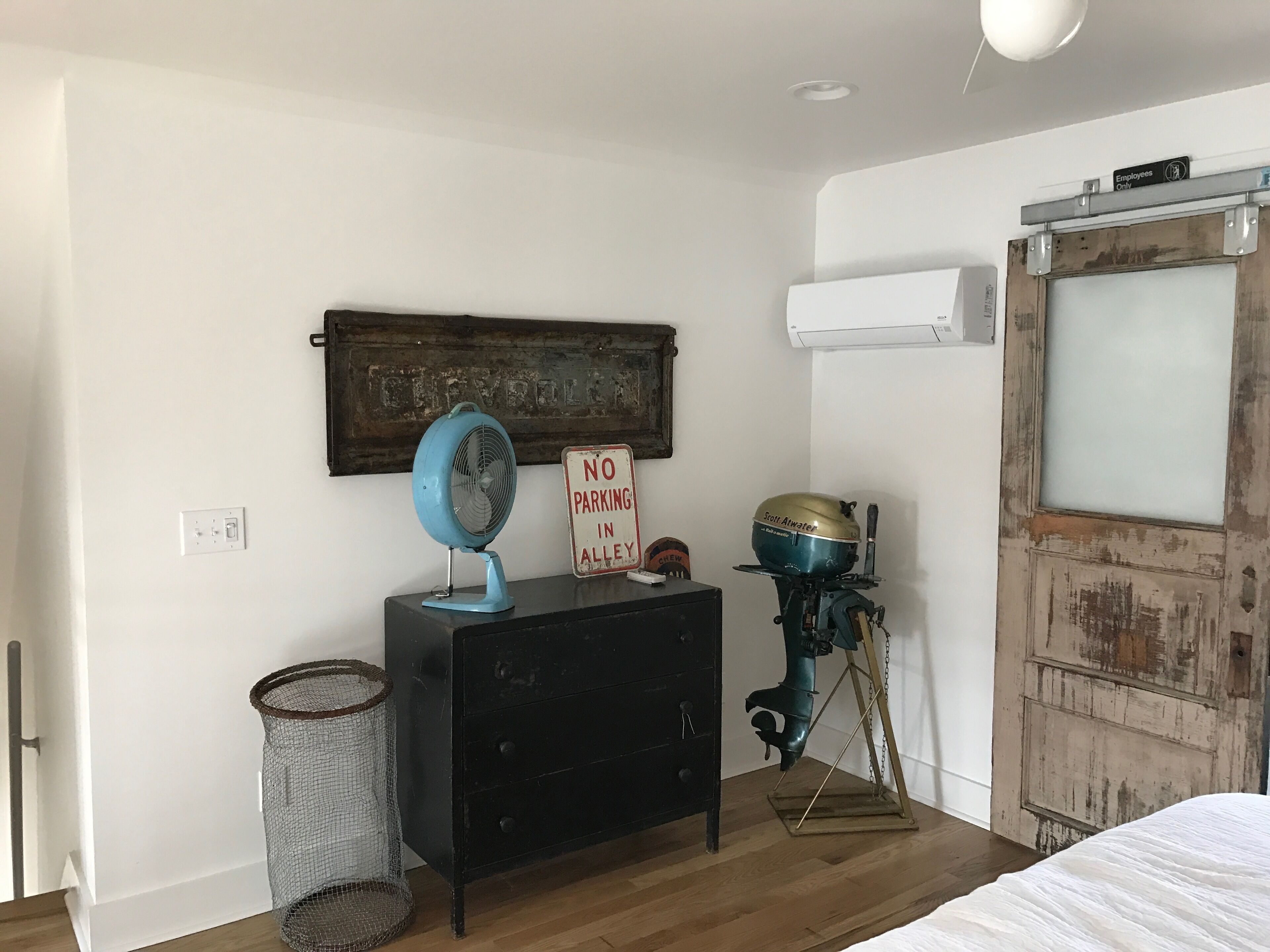 Une chambre avec un lit, une commode, un ventilateur ancien, un panneau indiquant qu’il est interdit de se garer, et une porte rustique.