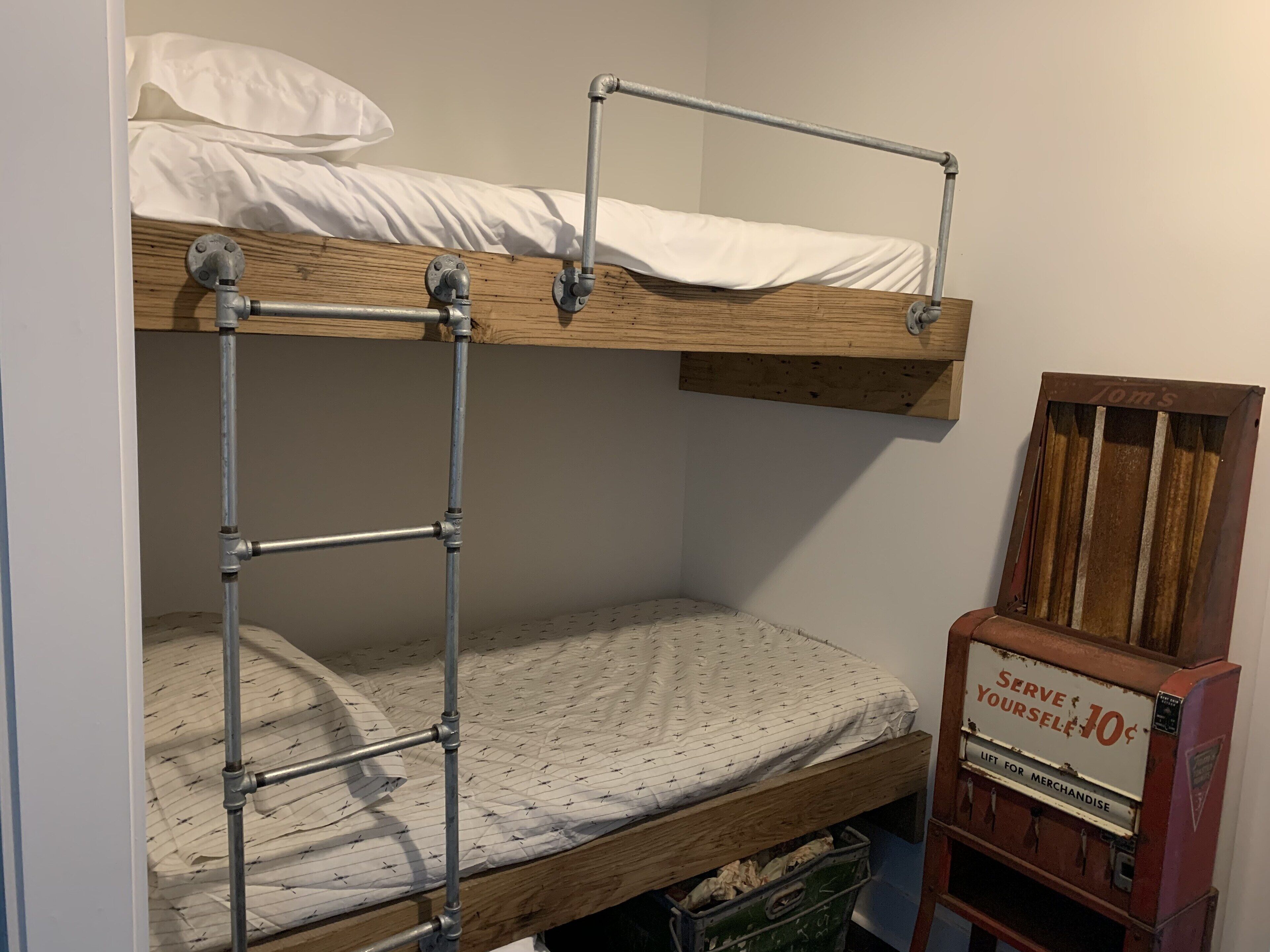 A bunk bed with a metal ladder, a wooden headboard, and a mattress with a patterned cover. There is a vintage vending machine with a sign that says "Serve Yourself 10¢" and a green storage bin under the bed.